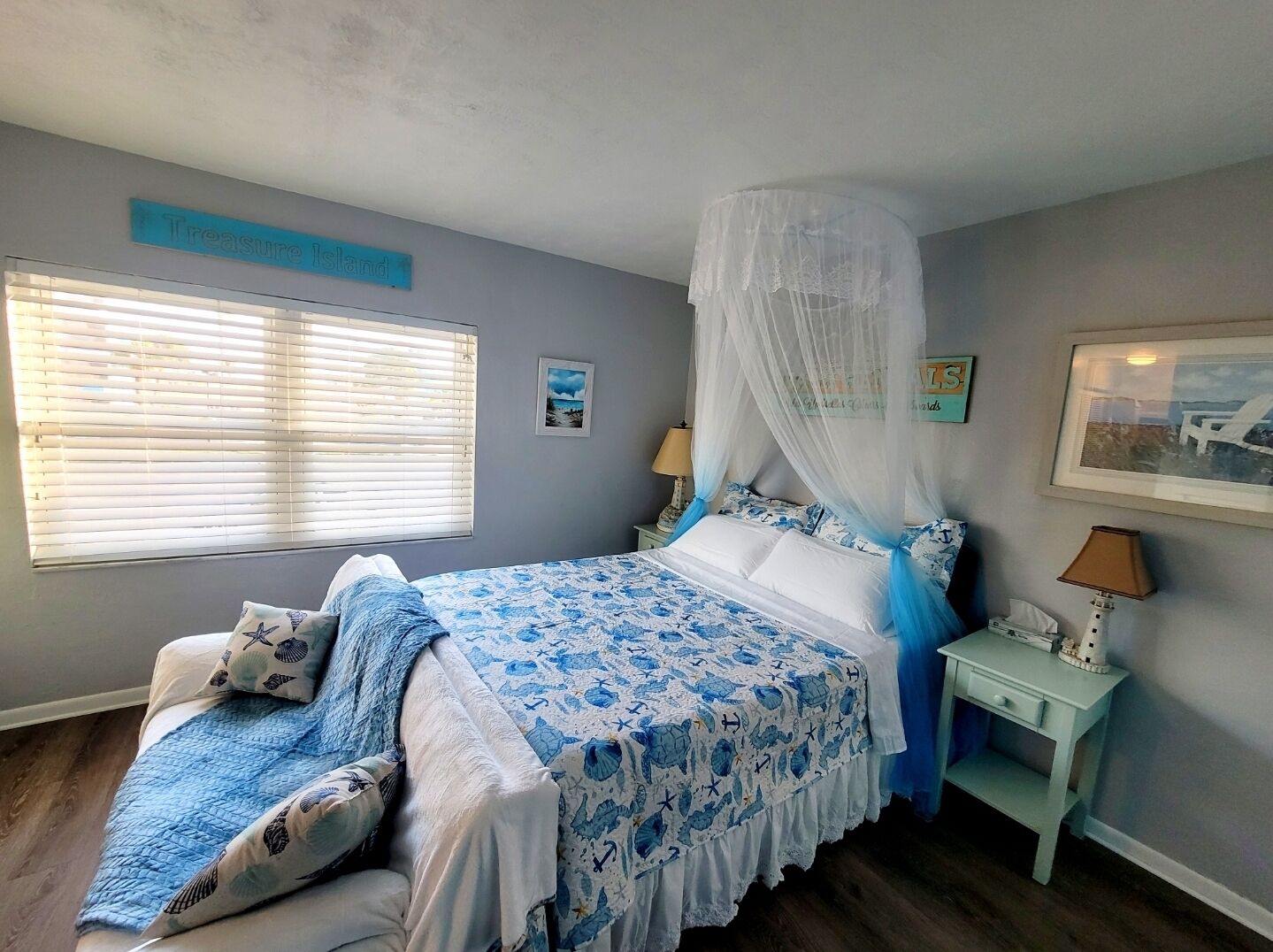 A bedroom with a bed under a canopy, a bedside table with a lamp, a framed picture on the wall, and a window with blinds.
