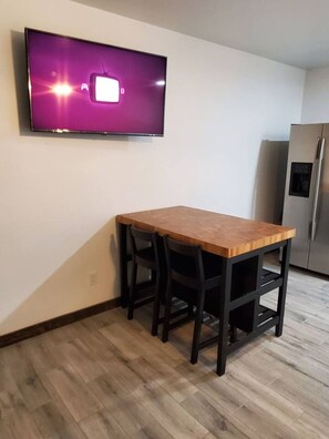 A small kitchen with a wooden table and chairs, a built-in TV, and a refrigerator.