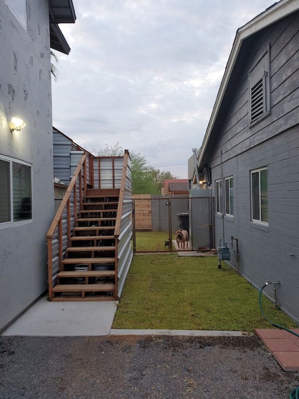 Un jardin avec un escalier en bois menant à une zone abritée, un enclos pour chiens délimité par une clôture et un tuyau d’arrosage posé par terre.