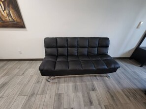 Un canapé en cuir noir, doté d’une structure en métal, est placé sur un plancher en bois dans une pièce aux murs blancs.