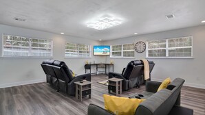 Amplia sala de estar con un sofá gris, dos sillones de masaje, una mesita y un televisor en la pared.