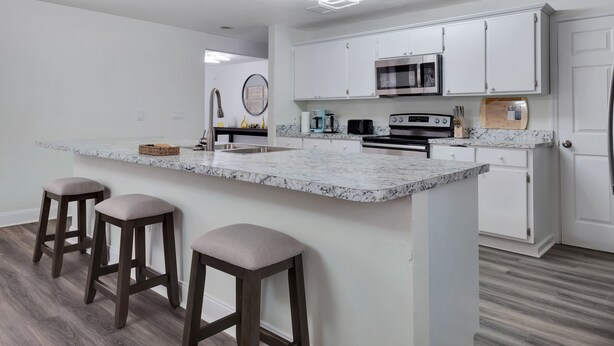 Una cocina moderna con armarios blancos, encimera de granito y horno empotrado. Hay tres taburetes de bar y una isla grande con taburetes.