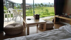 A bedroom with a bed, a table, two poufs, and a view of a green field and buildings. - Daxiagu Ligong Guoyuan Holiday Hotel (Ruyuan)