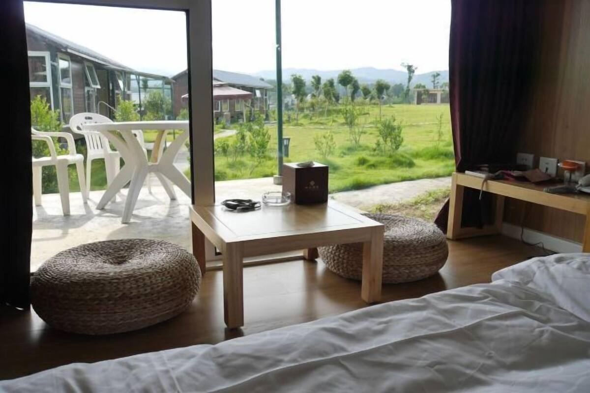 Ein Schlafzimmer mit einem Bett, einem Tisch, zwei Poufs und Blick auf ein grünes Feld sowie Gebäude.