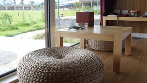 A room with a large window, a wooden table, a woven ottoman, and a view of a grassy area. - Daxiagu Ligong Guoyuan Holiday Hotel (Ruyuan)