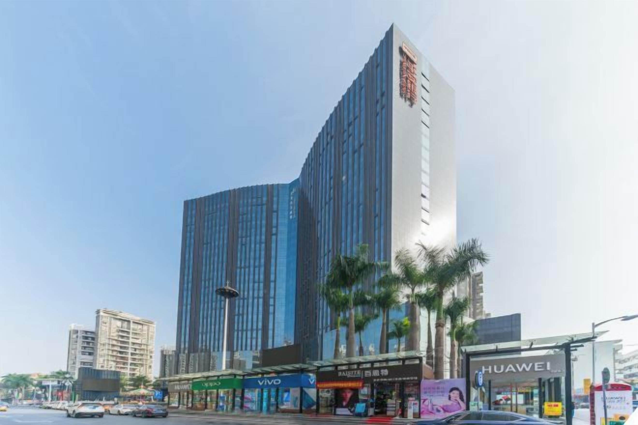 A modern high-rise building with a glass facade, surrounded by palm trees and a street with shops.