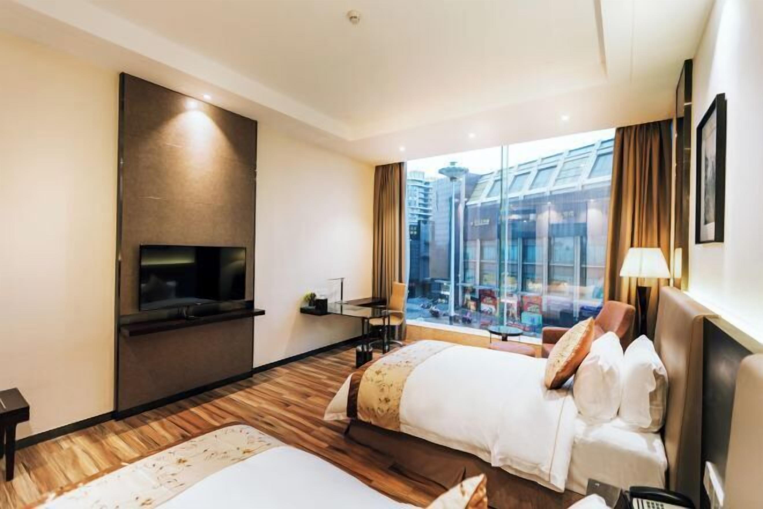 A hotel room with two beds, a TV, a desk, and a large window with a city view.