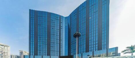 Un moderno edificio de gran altura con ventanas de vidrio reflectante, un reloj destacado y locales comerciales en la planta baja.