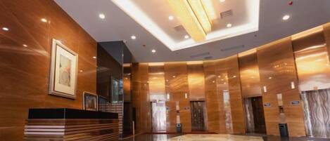 A modern hotel lobby with a reception desk, elevators, and a marble floor.