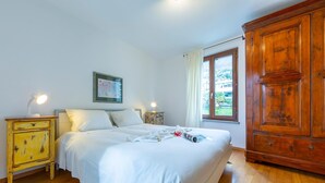Ein Schlafzimmer mit großem Holzschrank, einem Bett mit weißer Bettwäsche, einem Nachttisch mit Lampe und einem Fenster mit Vorhängen.