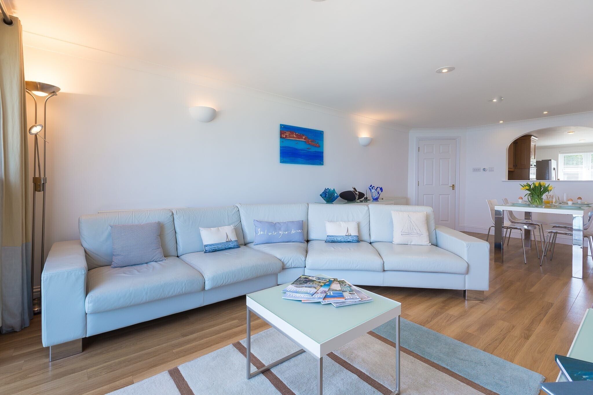 A modern living room with a light blue sectional sofa, a glass coffee table, and a dining area with a white table and chairs.