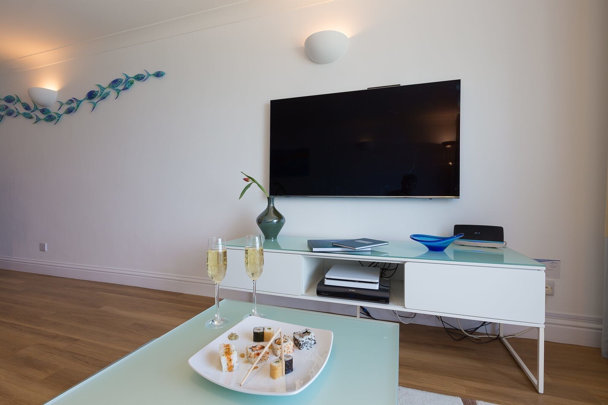 A modern living room with a flat-screen TV, a glass coffee table with a plate of sushi, two glasses of champagne, and a decorative wall piece.