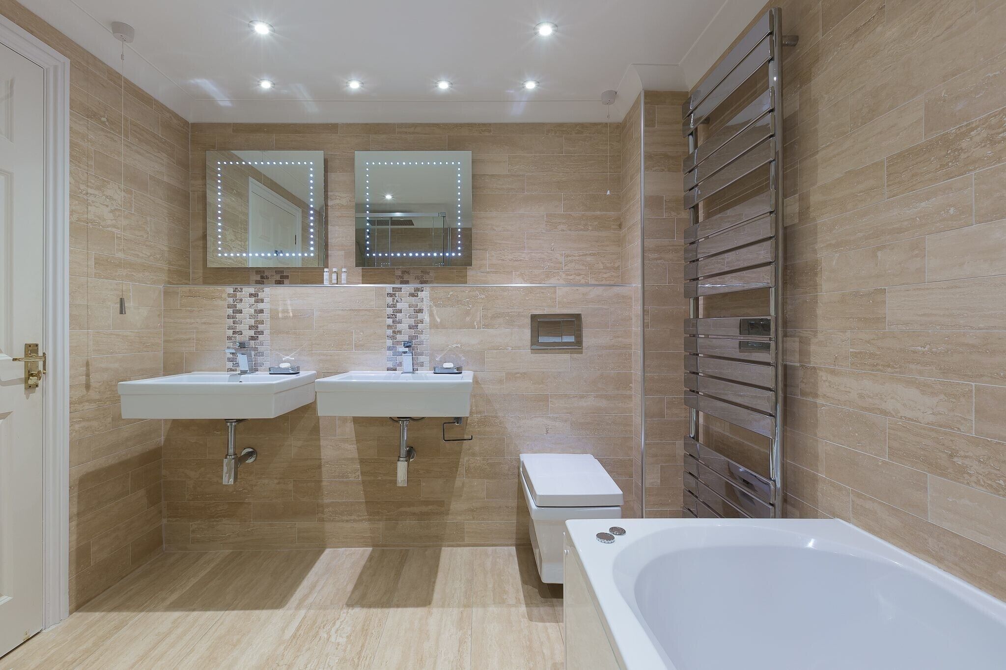 A modern bathroom with two sinks, a large mirror with lights, a heated towel rail, and a bathtub.