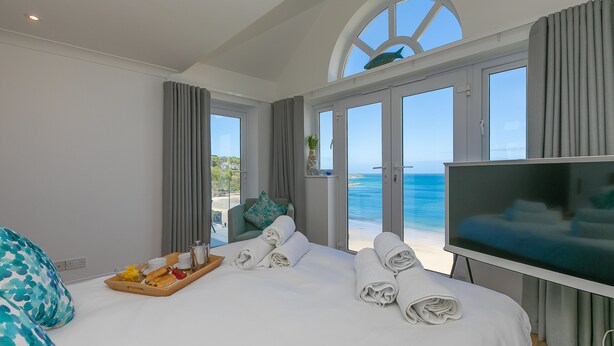 Une chambre d’hôtel avec un lit, une télévision, un plateau avec des articles pour le petit-déjeuner et une vue sur la mer par de grandes fenêtres.