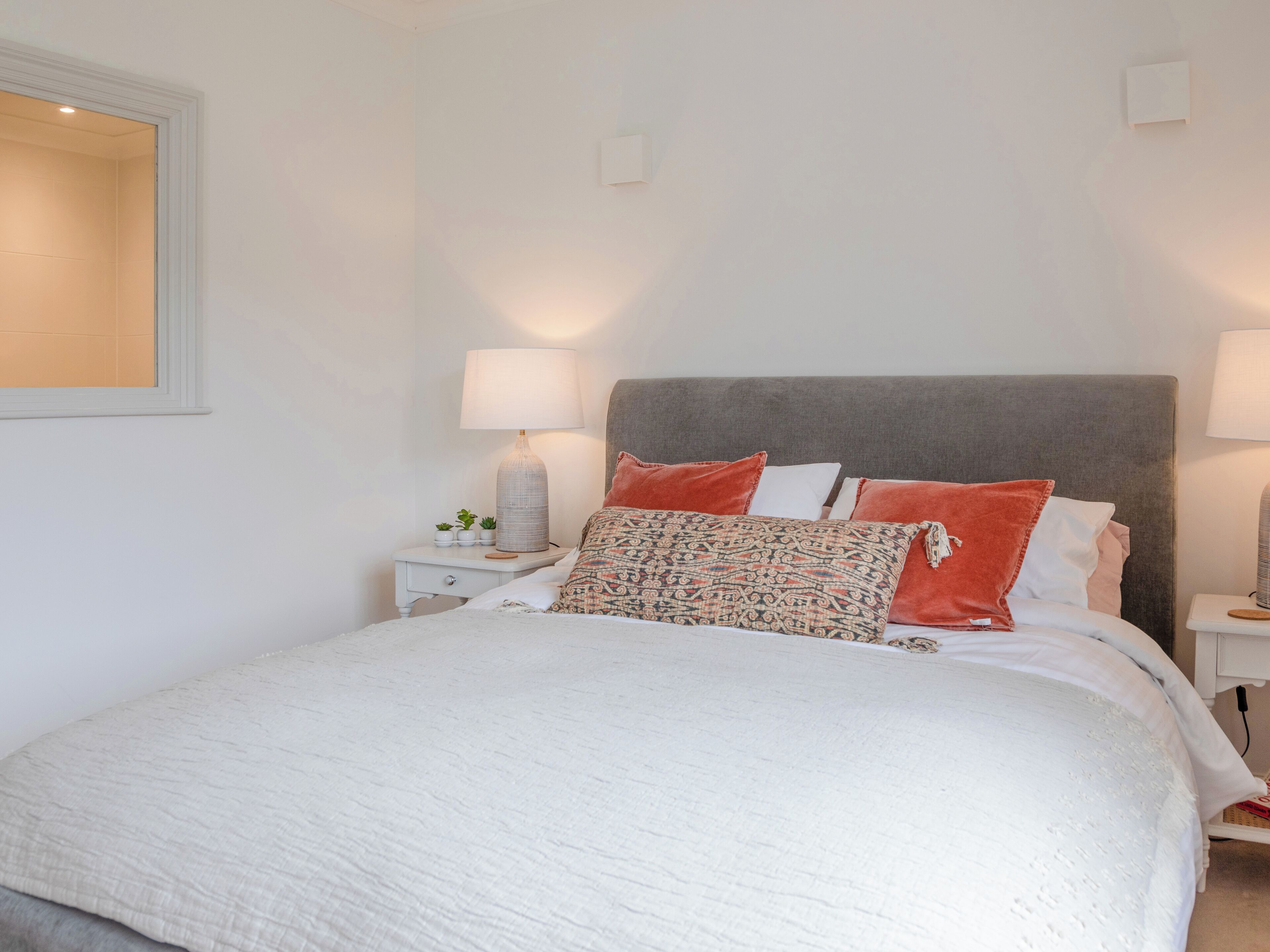 Una cama bien hecha con edredón blanco y varios almohadones, incluyendo diseños estampados y colores lisos. Hay dos mesitas de noche con lámparas y pequeños adornos.