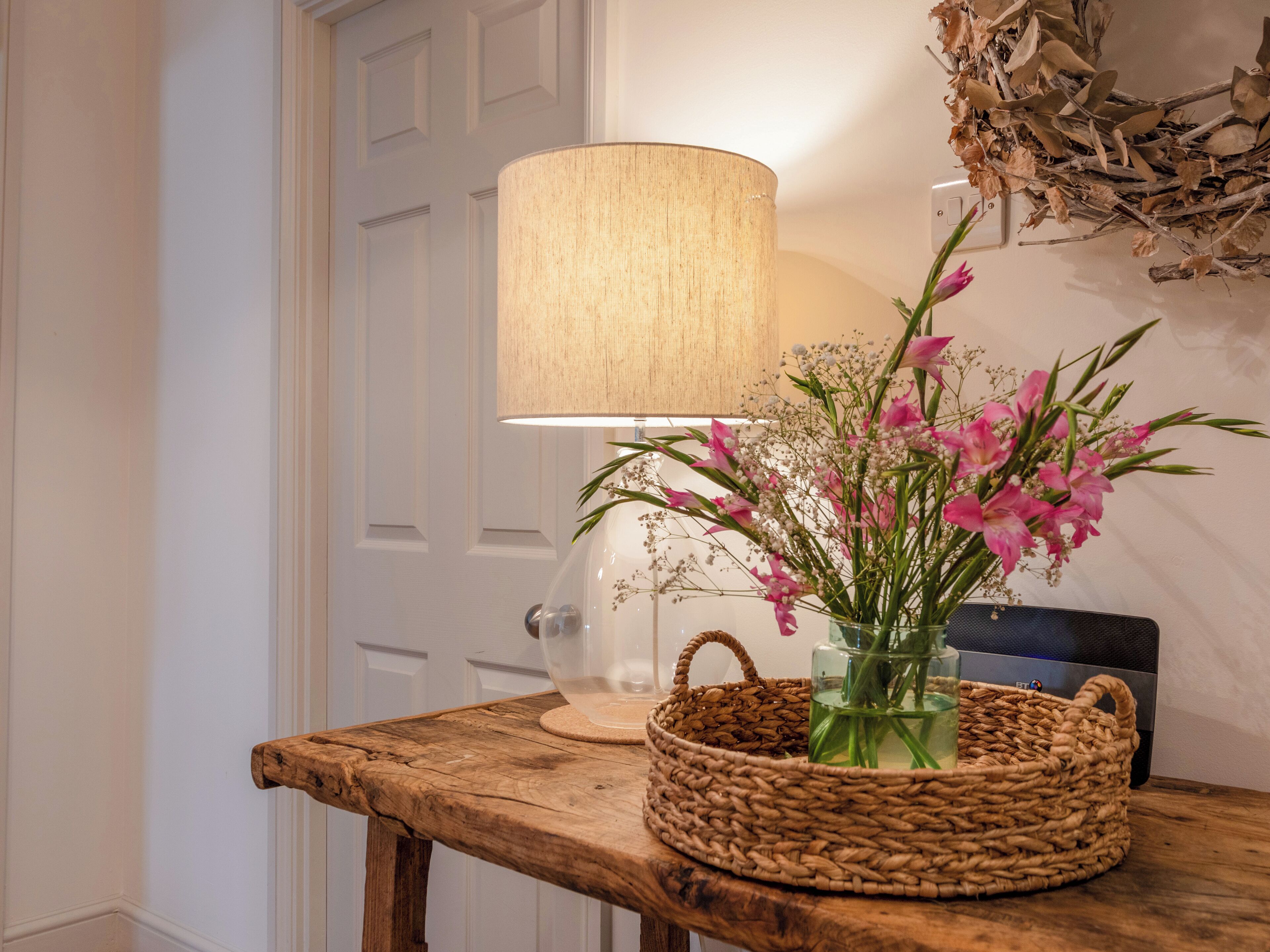 Ein Holztisch mit einem gewebten Korb, der eine Blumenvase und eine Lampe mit beigem Lampenschirm enthält.