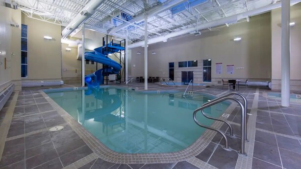 An indoor swimming pool with a blue slide, tiled flooring, and a high ceiling with visible structural beams. - Spacious Condo, Great Location & Mountain Views (Canmore)