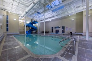 An indoor swimming pool with a blue slide, tiled flooring, and a high ceiling with visible structural beams. - Spacious Condo, Great Location & Mountain Views (Canmore)