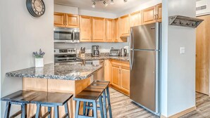A modern kitchen with wooden cabinets, stainless steel appliances, a granite countertop, and a built-in island with bar stools. - Spacious Condo, Great Location & Mountain Views (Canmore)