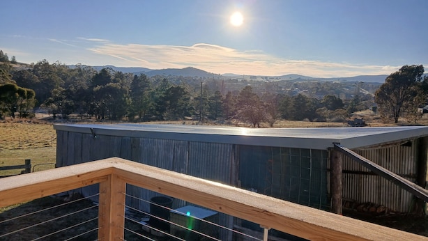 Une vue depuis une terrasse en bois dotée de rampes métalliques, donnant sur un paysage rural avec des arbres et des collines à l’horizon.
