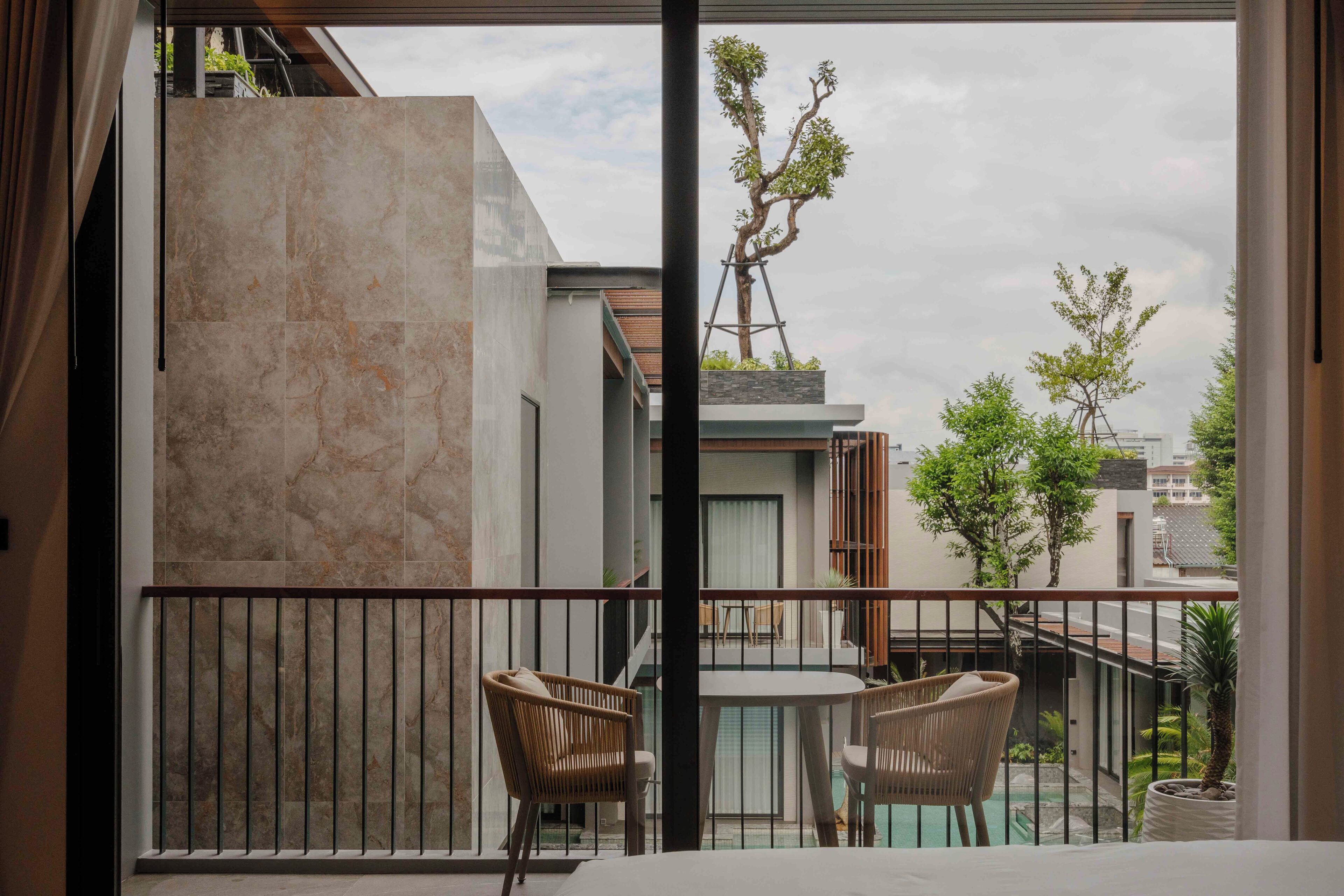 A balcony with two wicker chairs, a small table, and a view of a tree and buildings.