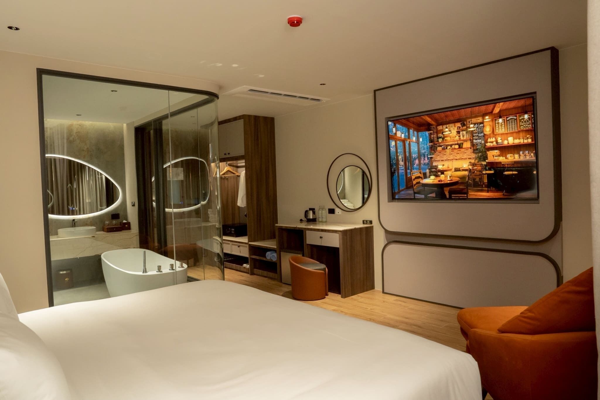 A modern hotel room with a large bed, a glass-enclosed bathroom, a wooden vanity, and a dining area visible through the glass.