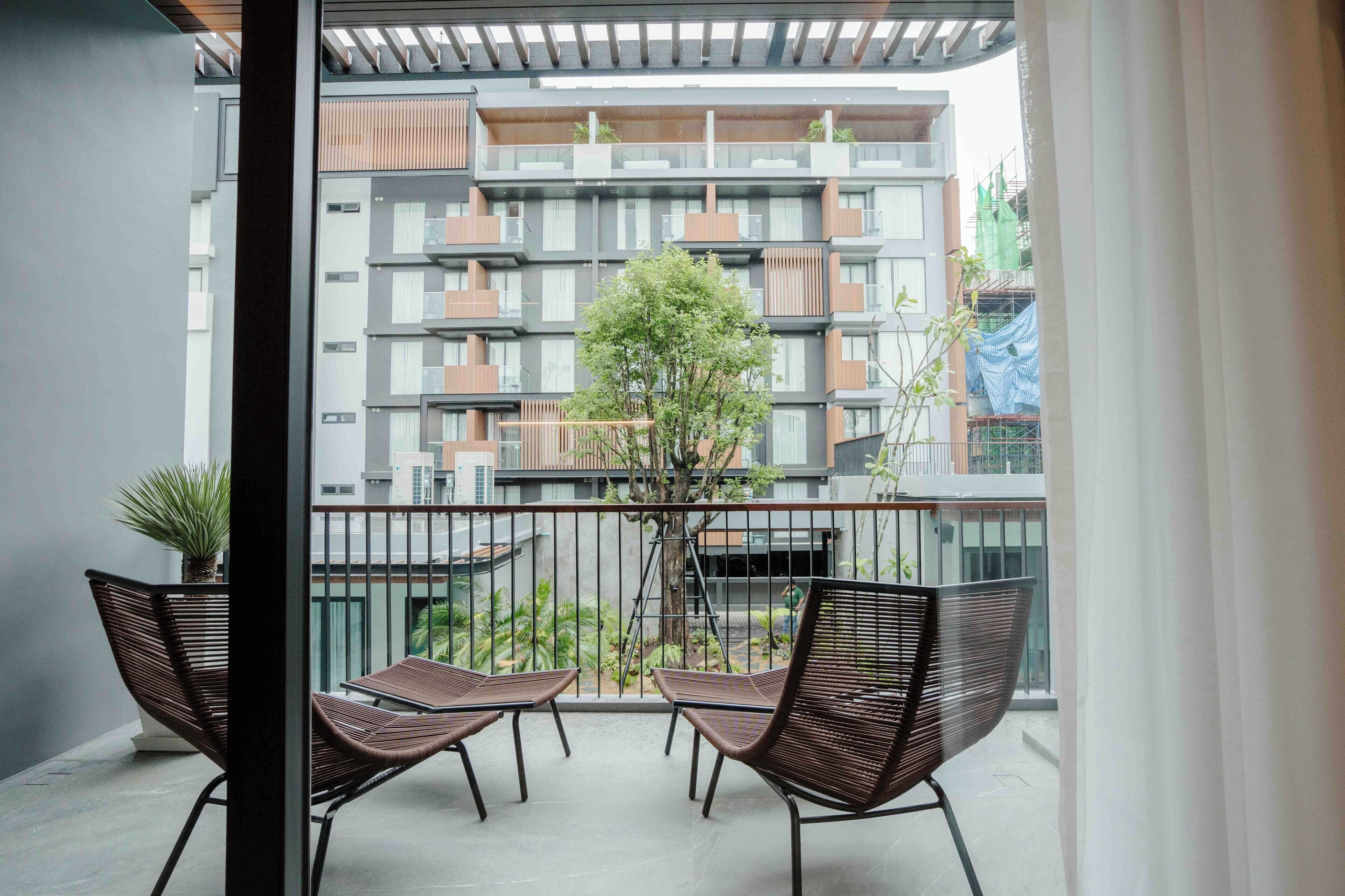 A balcony with two wicker chairs, a potted plant, and a view of a multi-story building with balconies.