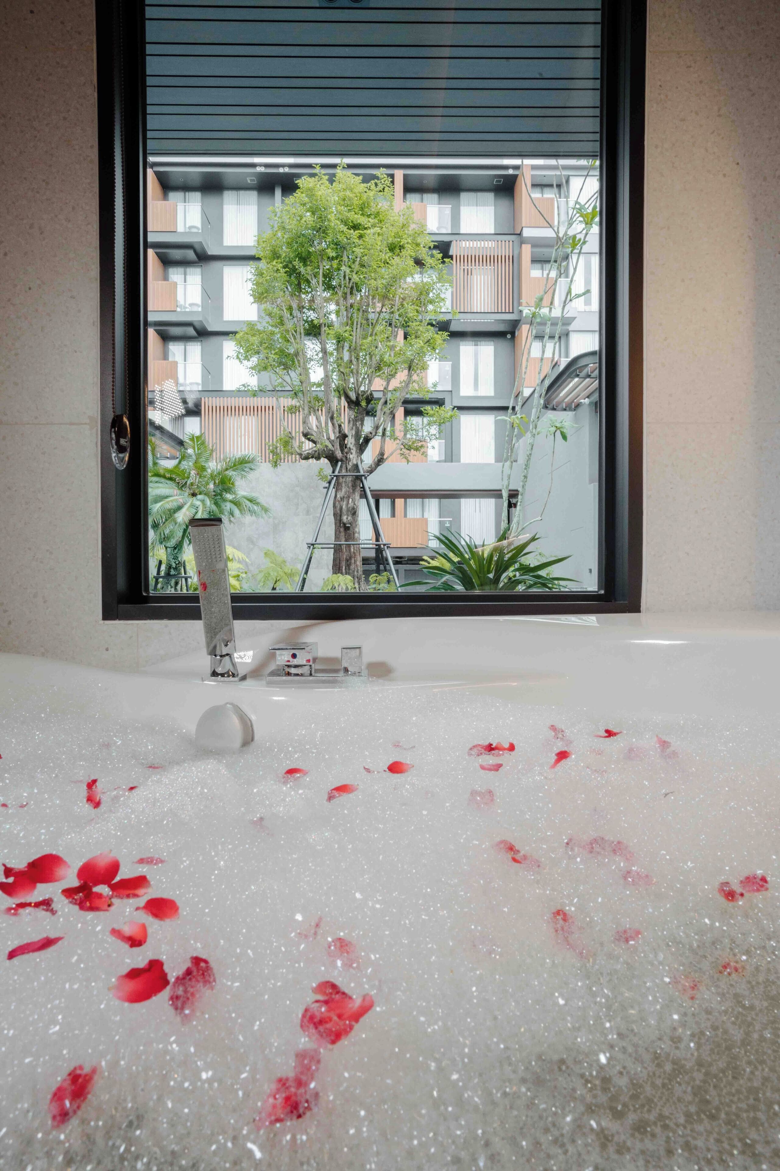 Une baignoire remplie de pétales de rose, une fenêtre et une vue sur un immeuble doté de balcons et de plantes.
