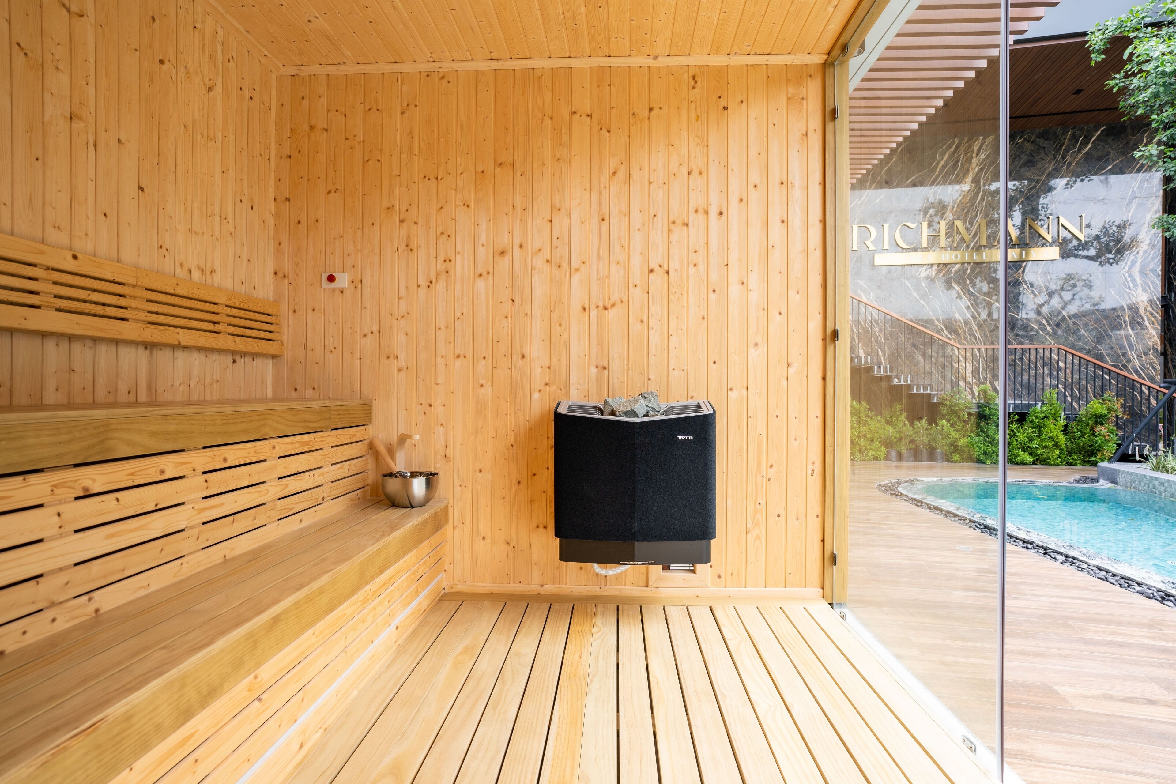 A wooden sauna with a bench, a black heater, and a glass door leading to a pool area.