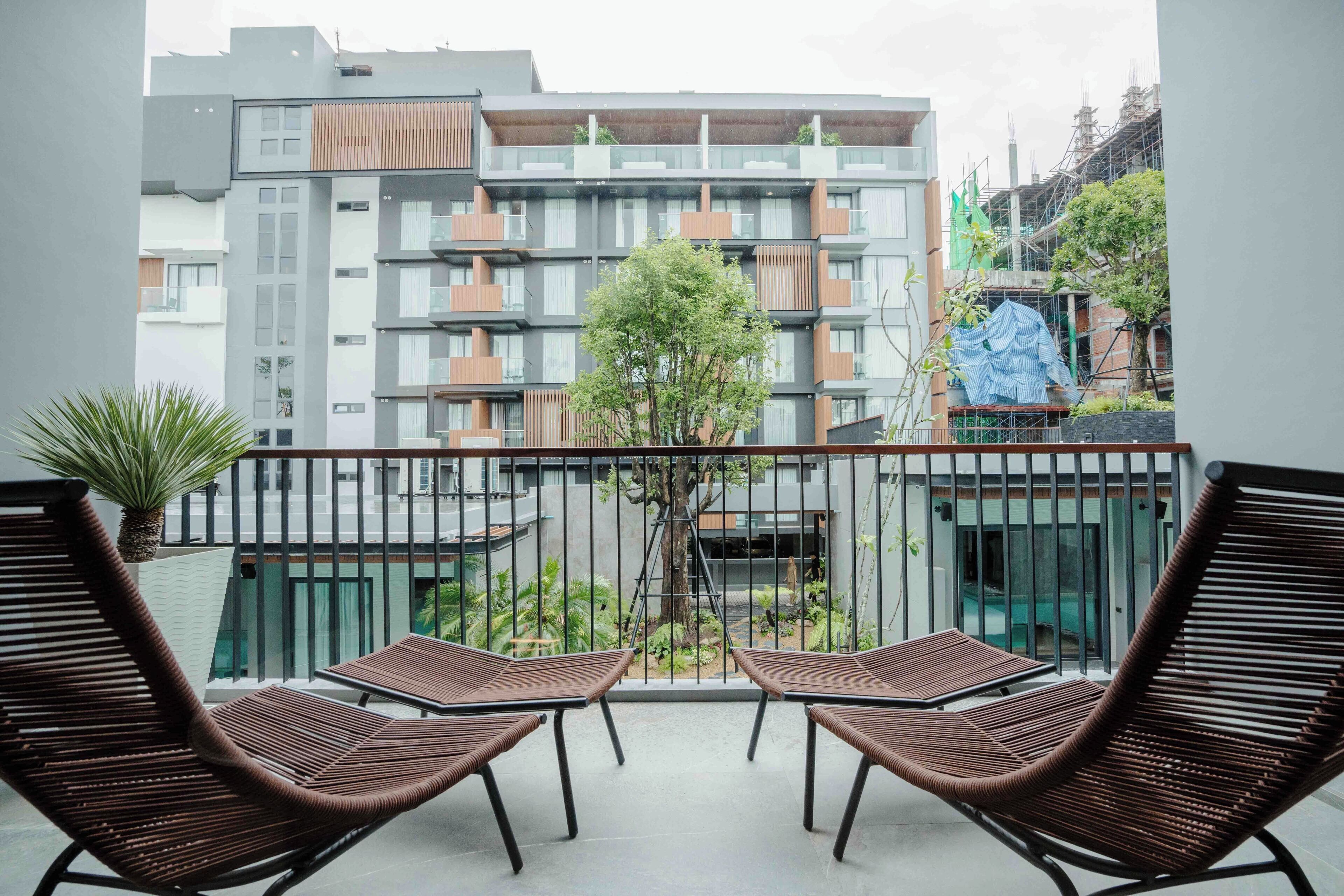 Un balcón con dos sillas de mimbre y una mesita, con vistas a un edificio moderno con varios balcones y un árbol en el centro.
