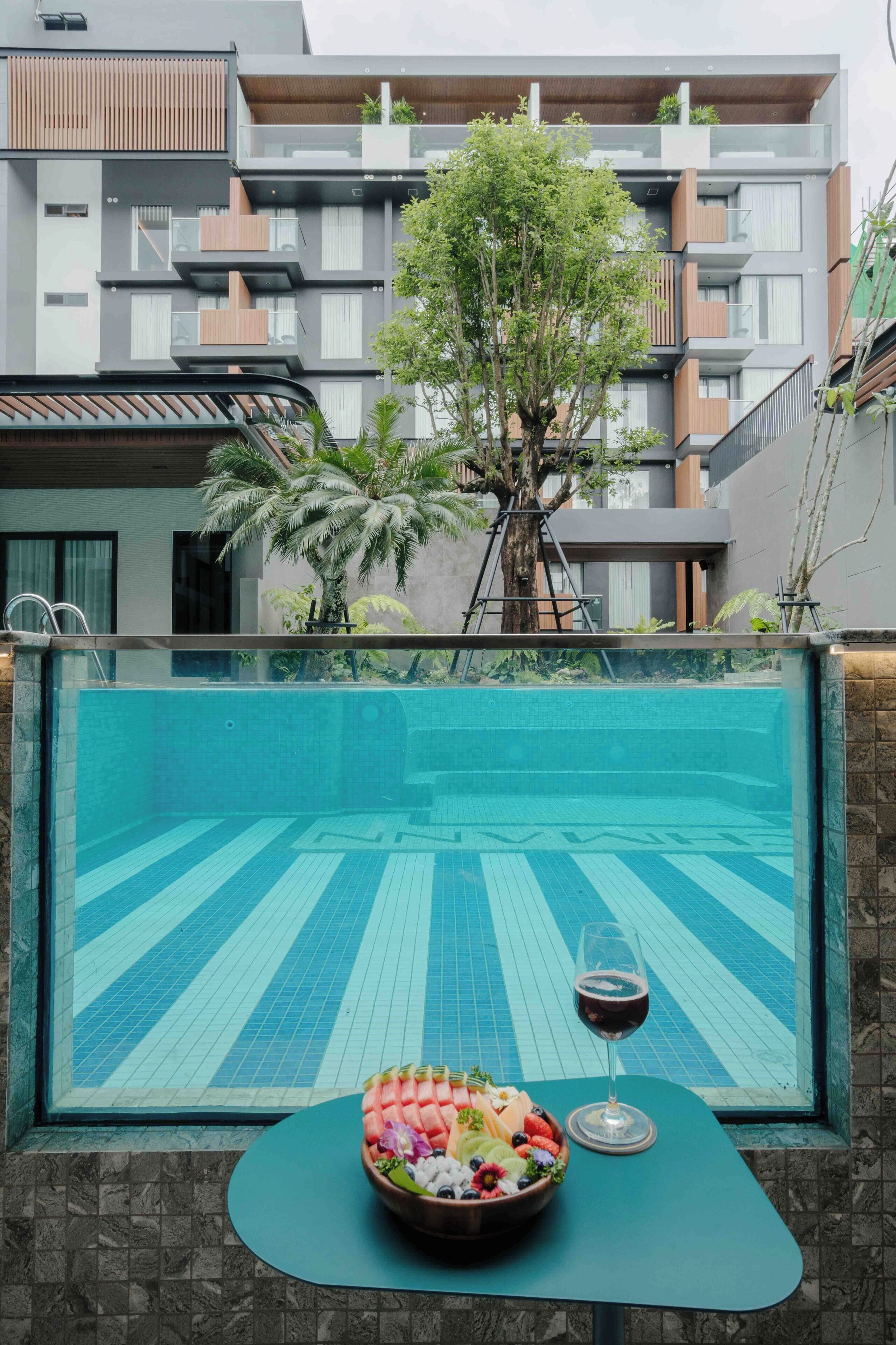 A swimming pool with a fruit bowl and wine glass on a table, surrounded by a modern building with balconies and a tree.