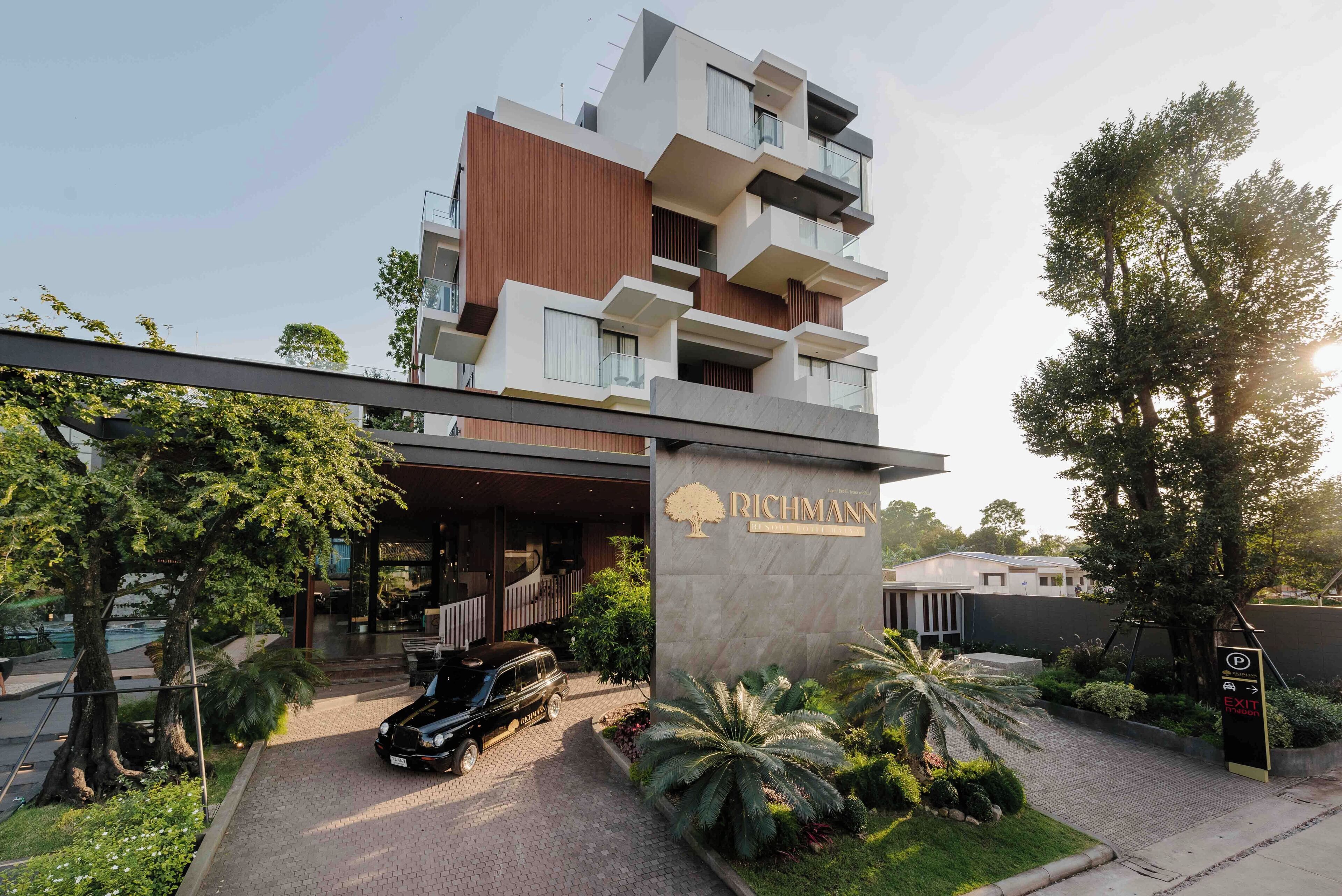 A modern multi-story building with a prominent entrance, a parking area, and lush landscaping.