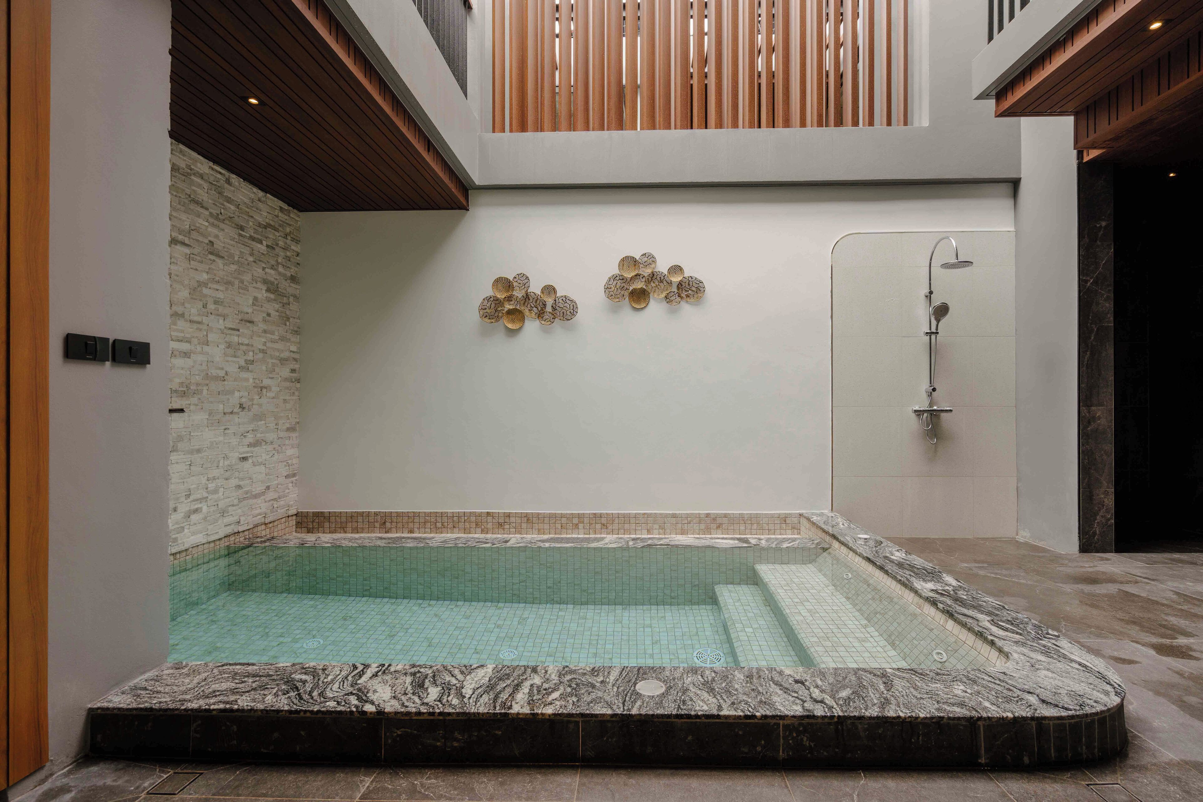 A modern indoor swimming pool with a stone border, a wooden ceiling, and a wall-mounted shower.