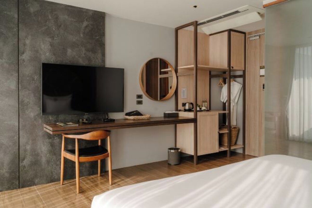 A modern hotel room with a large bed, a desk with a chair, a television, a wardrobe, and a small trash bin.