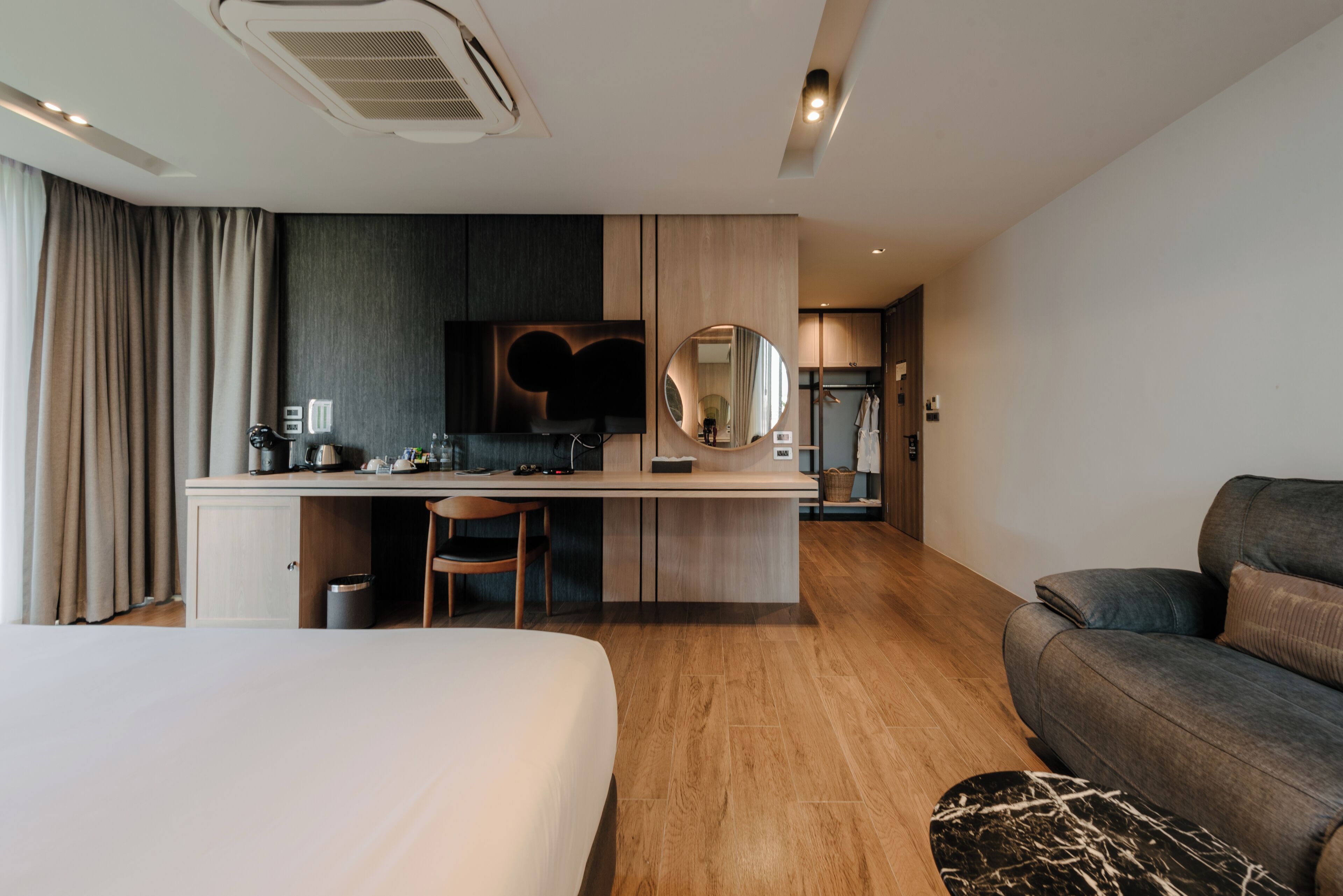 Una habitación de hotel moderna con cama, escritorio con silla, sofá, espejo redondo y armario.
