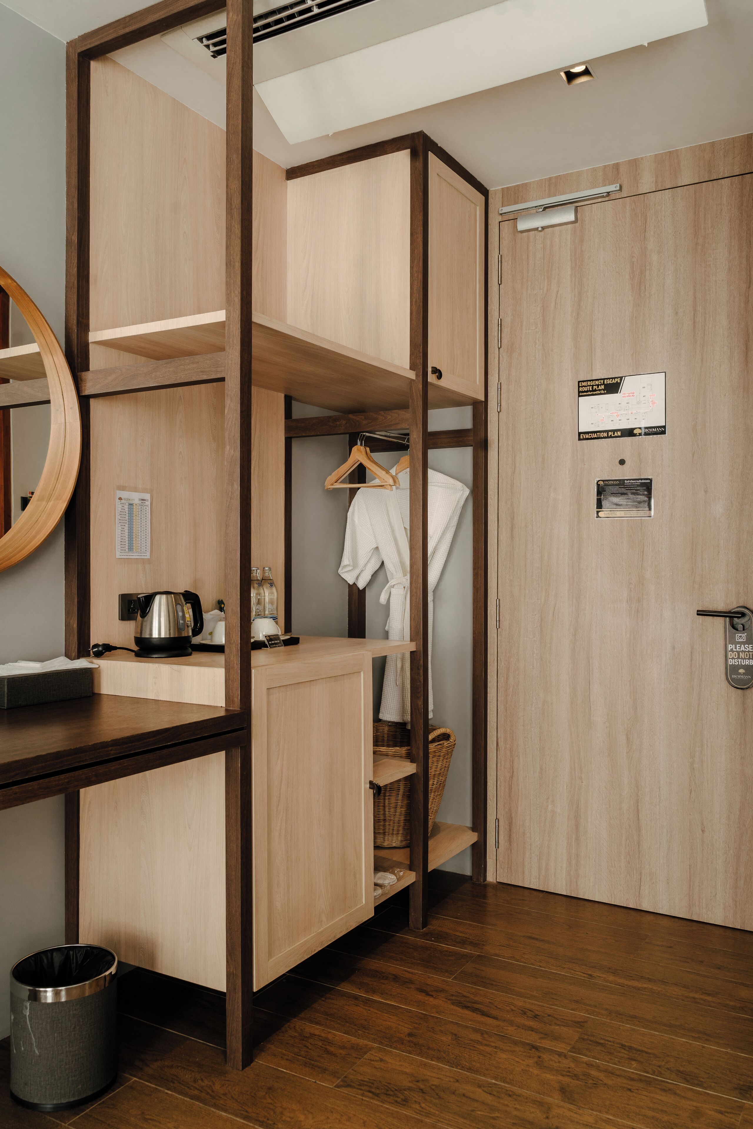 Une chambre d’hôtel avec une armoire en bois, un miroir, une petite table avec une bouilloire, une poubelle et une porte dotée d’une serrure et d’une pancarte.