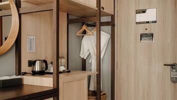 A hotel room with a wooden wardrobe, a mirror, a small table with a kettle, a trash bin, and a door with a lock and a sign.