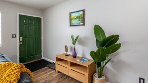Une pièce avec une porte verte, une console en bois sur laquelle sont posés un vase et un magazine, une grande plante et un tableau accroché au mur.