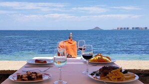 Mesa posta com pratos de comida e copos de vinho, com vista para um corpo de água e um horizonte urbano distante.
