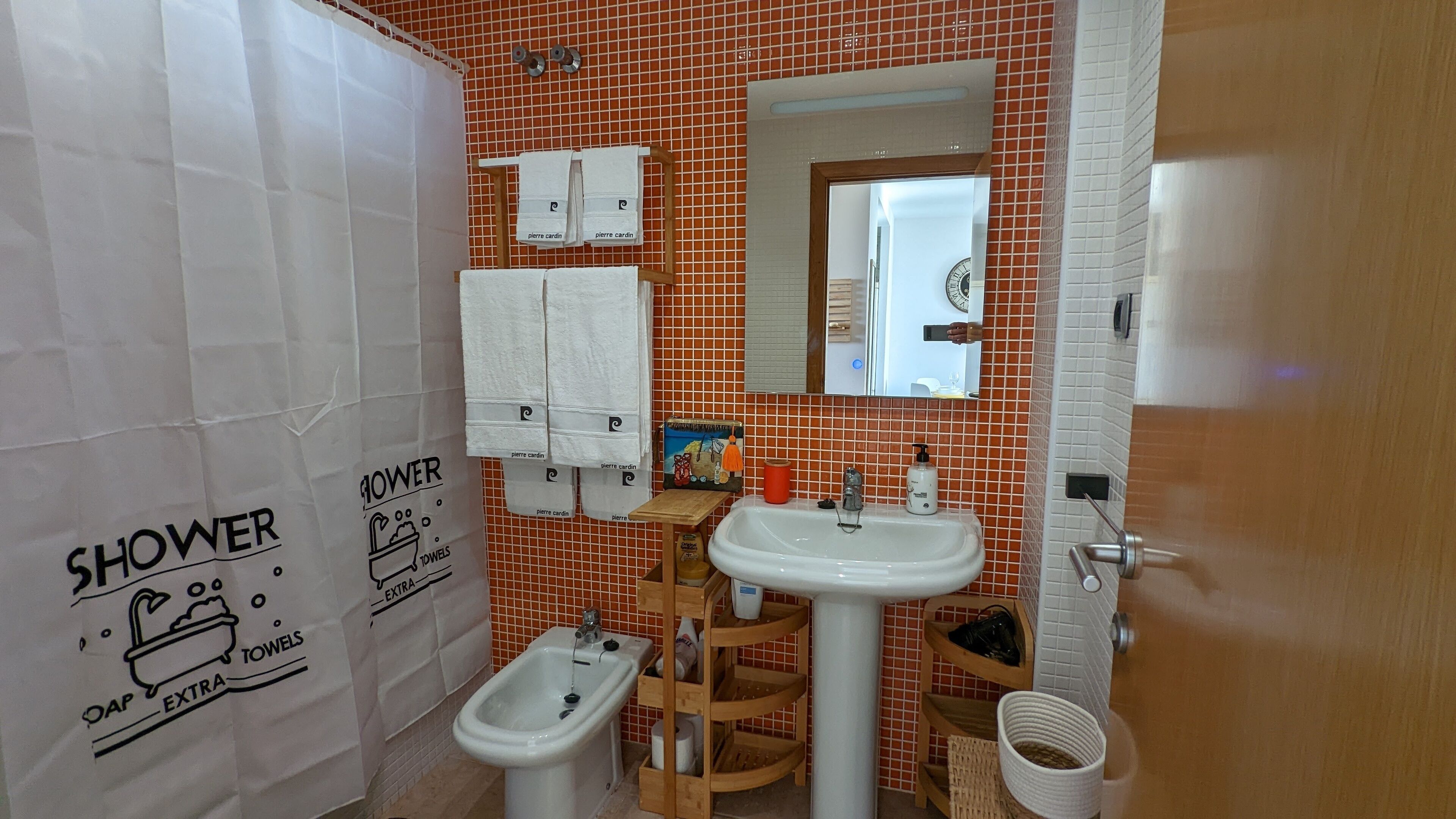 Un baño con ducha, lavamanos, bidé y espejo. Hay toallas colgadas en un perchero y una pequeña repisa con artículos de aseo.
