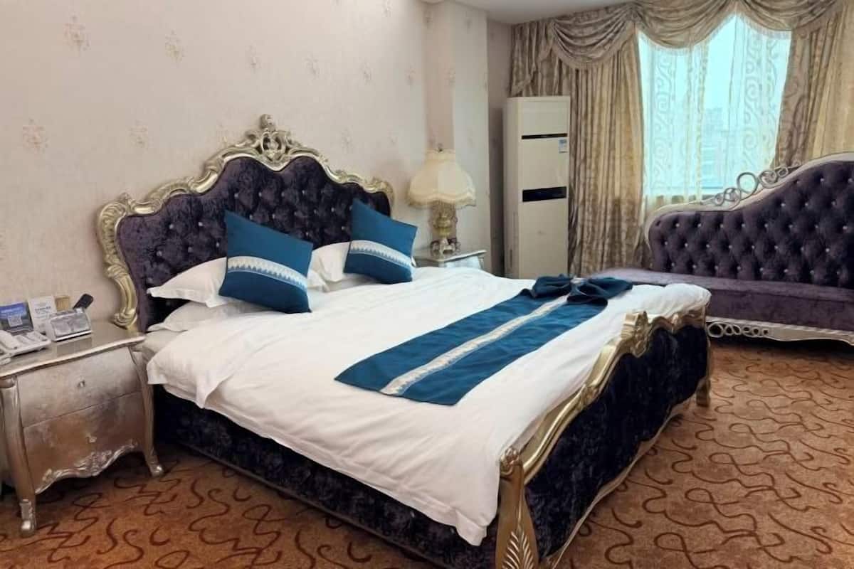 A hotel room with a large bed, a nightstand, a lamp, an air conditioning unit, and a window with curtains.