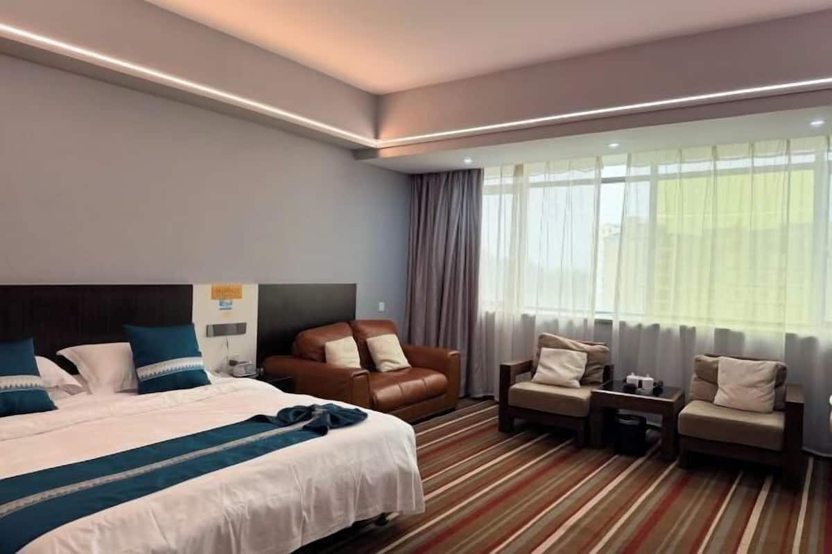 A hotel room with a bed, a brown leather sofa, two armchairs, a small table, and a window with curtains.