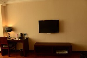 A hotel room with a flat-screen TV mounted on the wall, a wooden desk with a lamp, a chair, and a small table with a kettle and bottles. - Promenade Hotel (Pingnan)