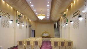 A banquet hall with rows of chairs, a stage with a golden backdrop, and a chandelier. - Hotel Virasat Palace By GRB (Mathura)