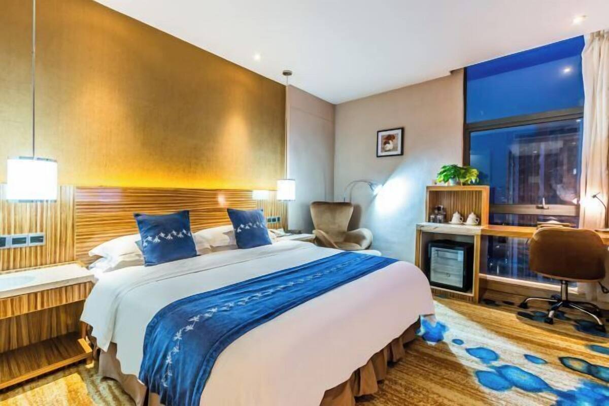 A hotel room with a bed, bedside table, lamp, chair, desk, and a large window.