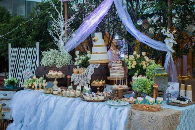 Una mesa de postres con diversos pasteles y repostería, rodeada de telas decorativas y arreglos florales.