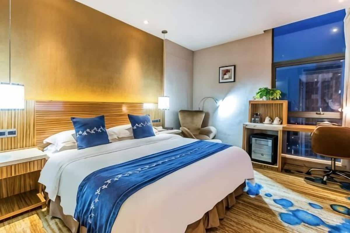 A hotel room with a bed, bedside tables, a desk, a chair, a desk lamp, a plant, and a window.