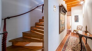 Una escalera de madera con barandilla de cuerda, una zona de comedor con mesa y sillas, y una alfombra estampada en el suelo.
