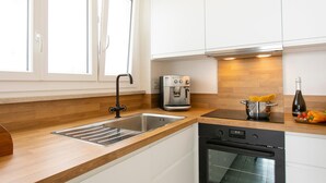Una cocina moderna con gabinetes blancos, un lavamanos de acero inoxidable, una cafetera, una estufa y una ventana con persianas.