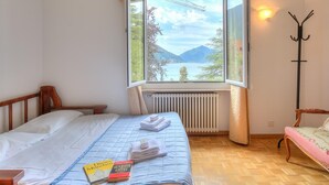 Una habitación con cama, una silla, una ventana con vistas a montañas y un lago, y un radiador.